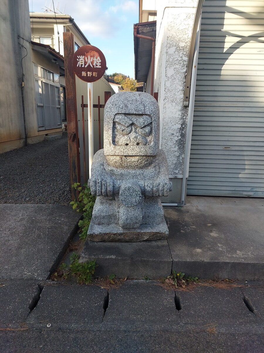 A traditional stone guardian figure with a marker sign stands outside a building in Iino-machi, Fukushima
